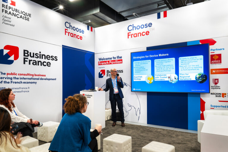 image of the CES 2025 Business France pavilion inside the trade show hall, showing a specialty area sectioned off by specialty walls to showcase a French business with a theater space, a representative is giving a presentation with a display screen for a gathered crowd.