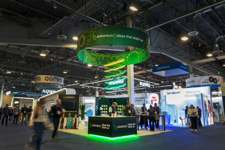 Image of Solventum's booth at CES 2025, from the trade show walkway, there is a large hanging sign with neon clouds descending from it to help communicate the multi-layered offerings of the company, below it there is a custom built counter to greet attendees