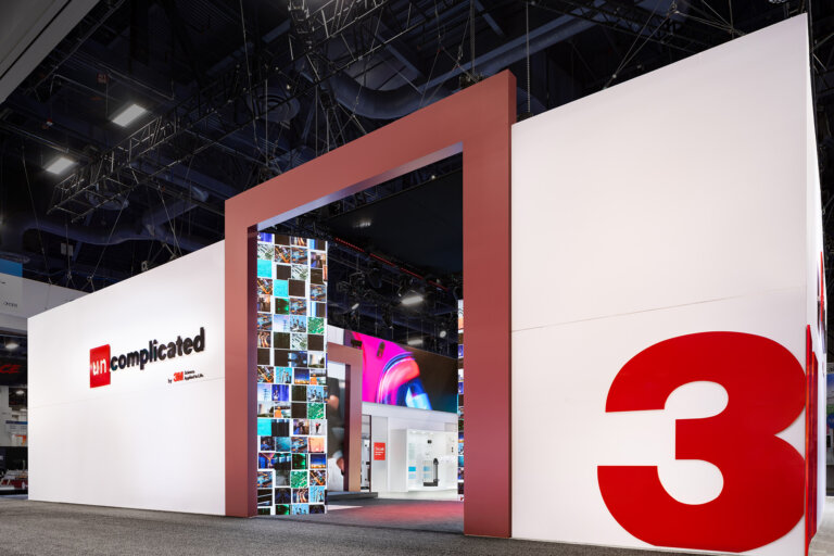 Image of the 3M booth at CES 2025, which is taken from the walkway. There are wall white walls in a box shape with the 3M logo on the outside, there is a large opening that is outlined in red, showcasing the inside of the booth space, with LED screens.
