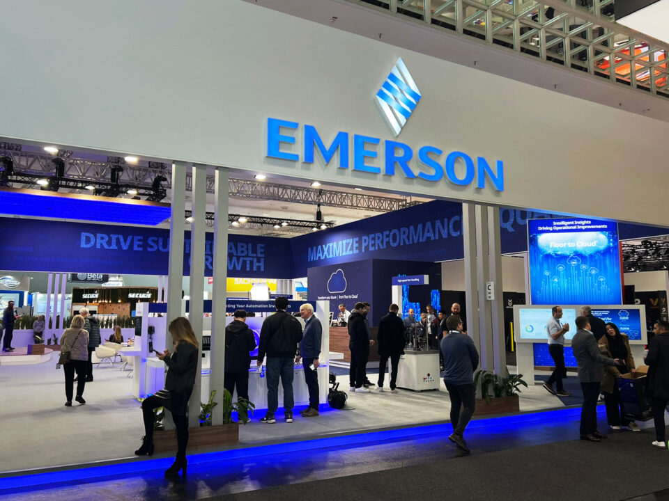 An image from the Emerson booth at the 2025 Hannover Messe trade show. This image showcases a wider view of the trade show display and exhibit space including people entering the trade show exhibit space below signage with the brand's logo.