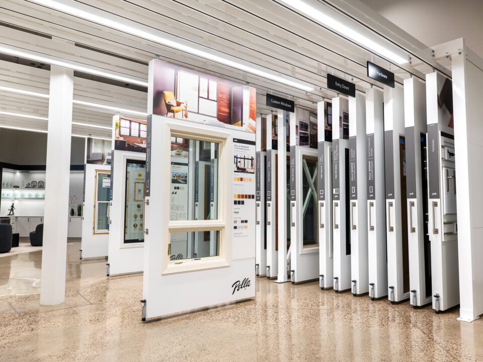 Image from Pella Northland HQ, featuring Pella displays that are housed in the showroom space of the headquarters. These are custom built displays for Pella doors and windows, that are neatly hung from the ceiling next to each other and are able to roll out to view the product and information on offerings and customizations. There is a seating area for client meetings in the background.