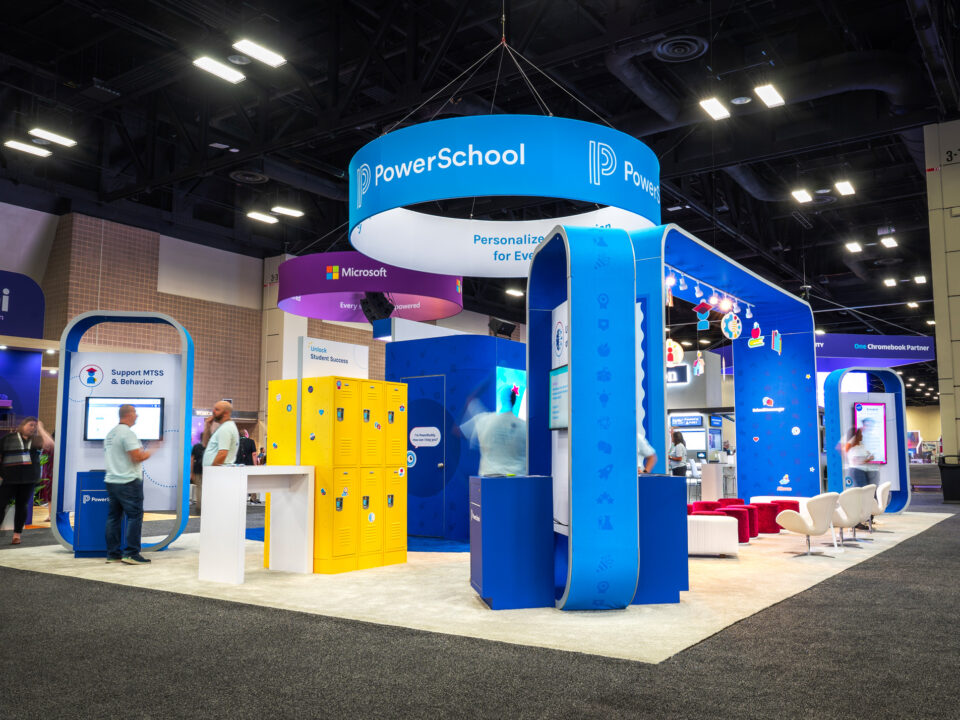 2025 PowerSchool ISTE overview of the trade show booth space. Their exhibit features a hanging ceiling sign with their logo on it. There are a variety of custom kiosks surrounding the space which have monitors on them. In the center you can see the theater space within the trade show exhibit, which has an arch over the attendees with custom graphics promoting the company. In the theater space there is a large LED screen and a variety of seating, including stools and chairs. On the left side of the image there is a custom giveaway activation which uses yellow lockers that trade show attendees received combinations to.