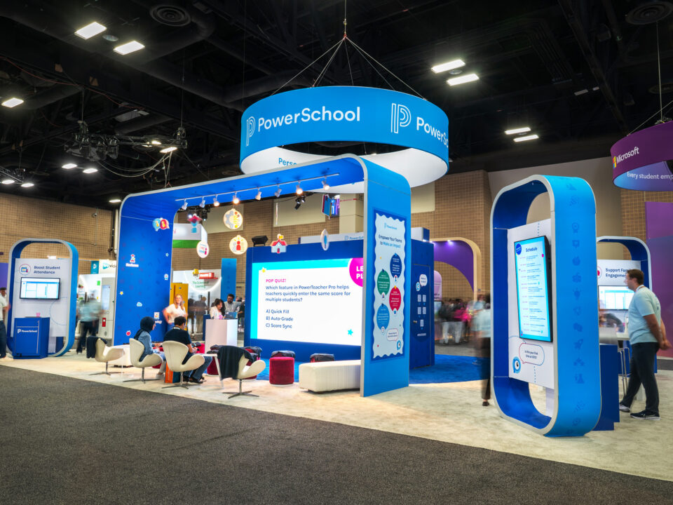 2025 PowerSchool ISTE overview of the trade show booth space. Their exhibit features a hanging ceiling sign with their logo on it. There are a variety of custom kiosks surrounding the space which have monitors on them. In the center you can see the theater space within the trade show exhibit, which has an arch over the attendees with custom graphics promoting the company. In the theater space there is a large LED screen and a variety of seating, including stools and chairs. There is a trivia game on the screen.