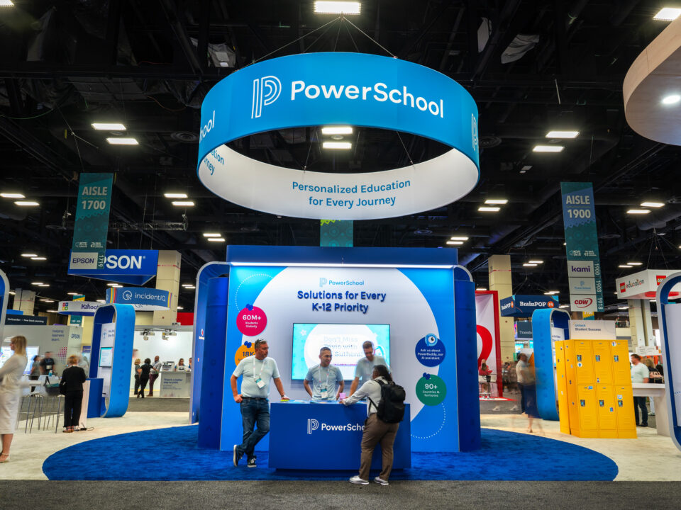 2025 PowerSchool ISTE overview of the trade show booth space. Their exhibit features a hanging ceiling sign with their logo on it. A welcome counter where employees greet a trade show attendee, that features a large monitor and customized tablecloth. There are a variety of custom kiosks surrounding the space which have monitors on them. In the right side of the booth you can see yellow lockers used for a customized giveaway activation on the trade show floor.