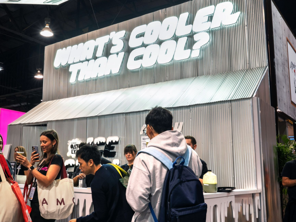 Image of Oatly's 20x20 booth at Expo West Natural Foods Expo 2025 featuring a giant sign on a metal background that says "what's cooler than cool?" and a barista bar serving Oatly iced coffee drinks. An attendee stands in front taking a photo of her drink.