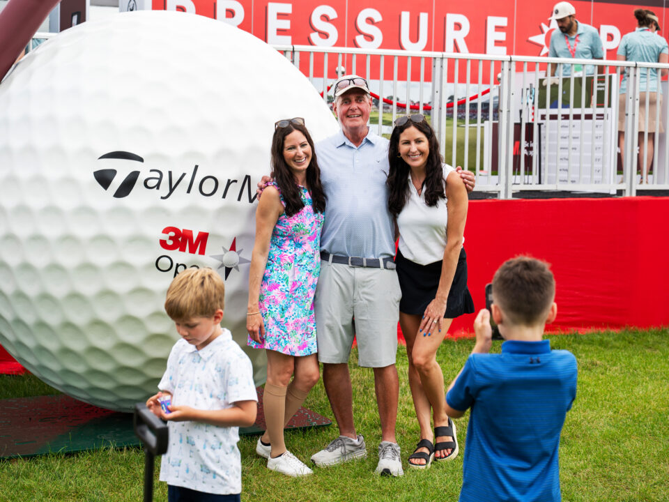 Image of a statue of a large golf ball and tee with the hashtag #golfthatmatters on them as well as the TaylorMade logo and the logo for the 3M Open. There is a family posing for a picture in front of it.