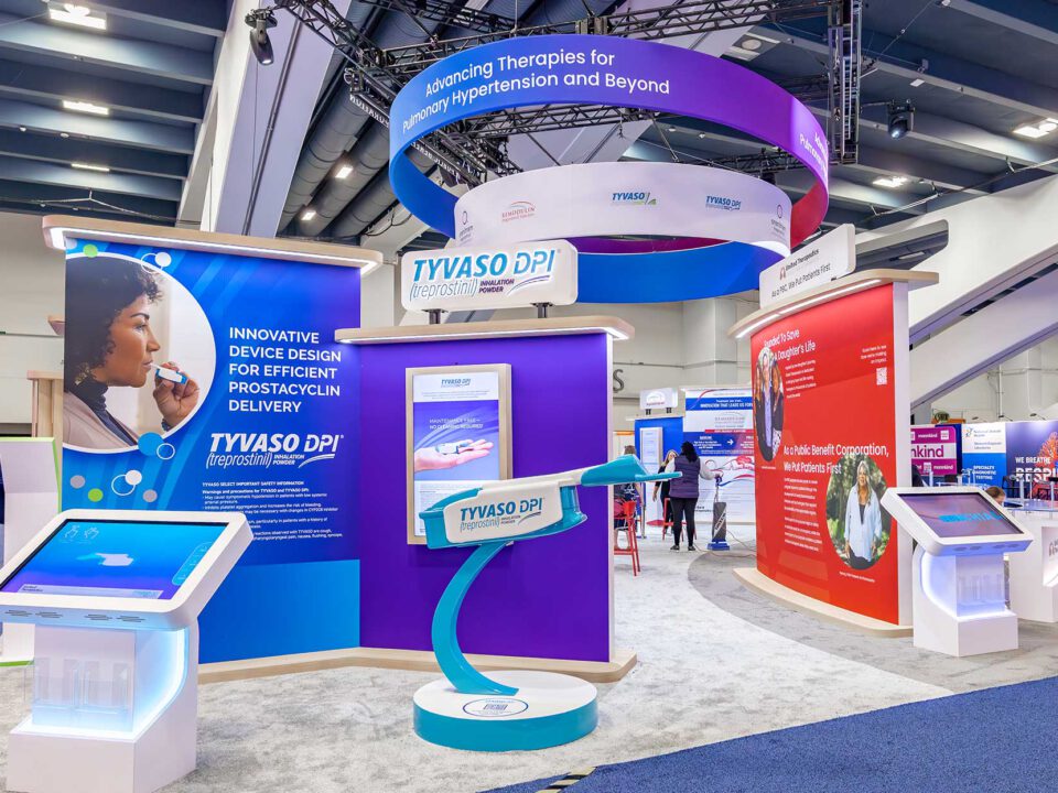 image of a booth designed for United Therapeutics, there are hanging signs from the ceiling, a few standing walls with information on products, a couple of interactive displays, and a large model of TYVASO DPI.